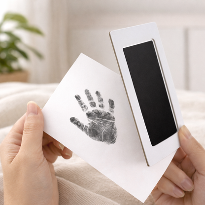 Baby handprint on white card made with an ink pad kit, held in hands against a soft, neutral Scandinavian-style interior background.